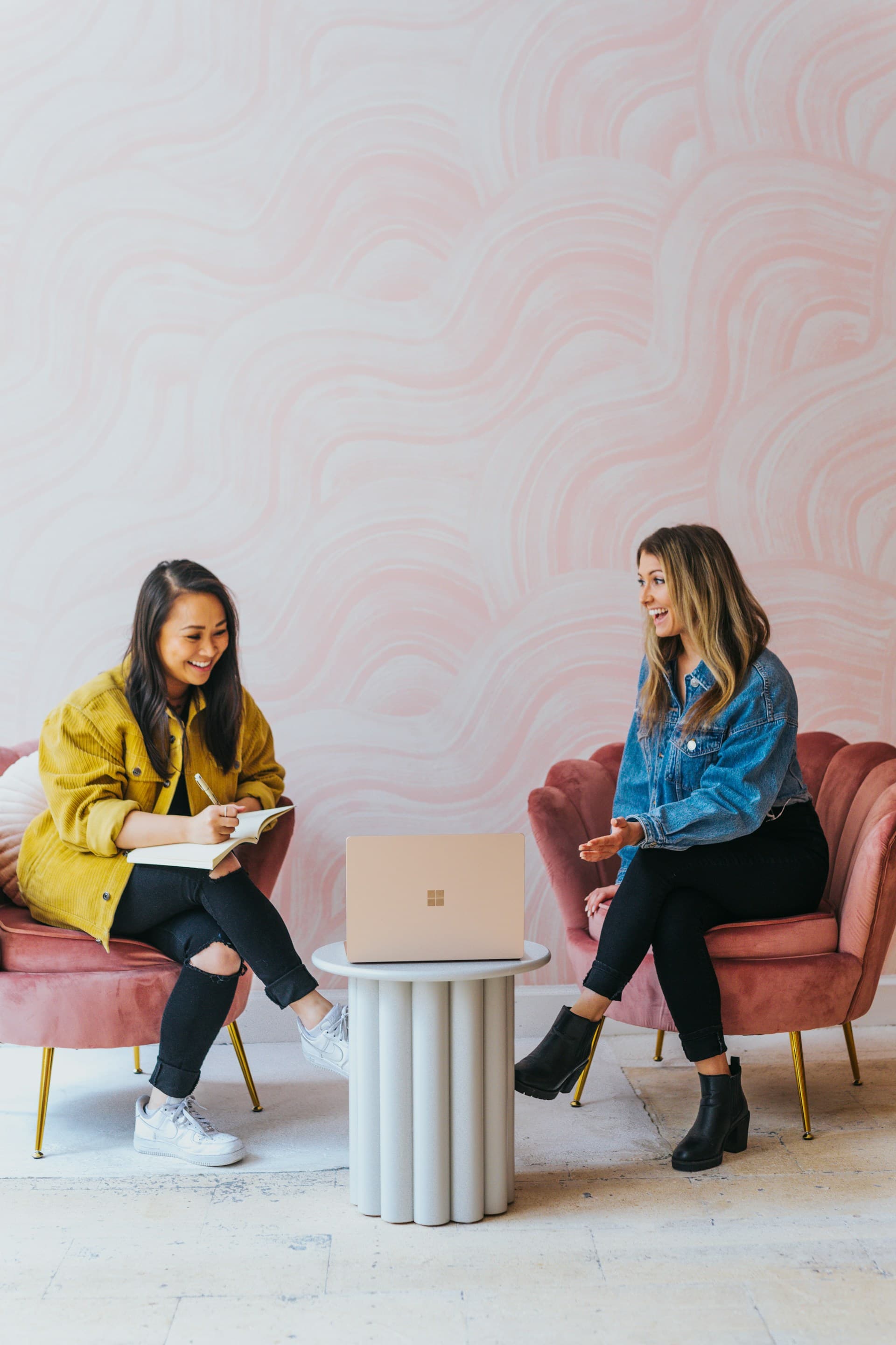 Two women sitting in comfy pink chairs collaborating over a shared laptop